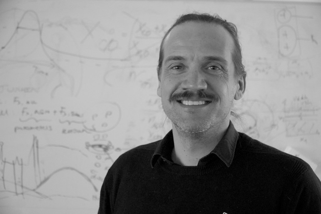 Portrait of Dr. Daniel Angerhausen standing in front of whiteboard showing equations and graphs