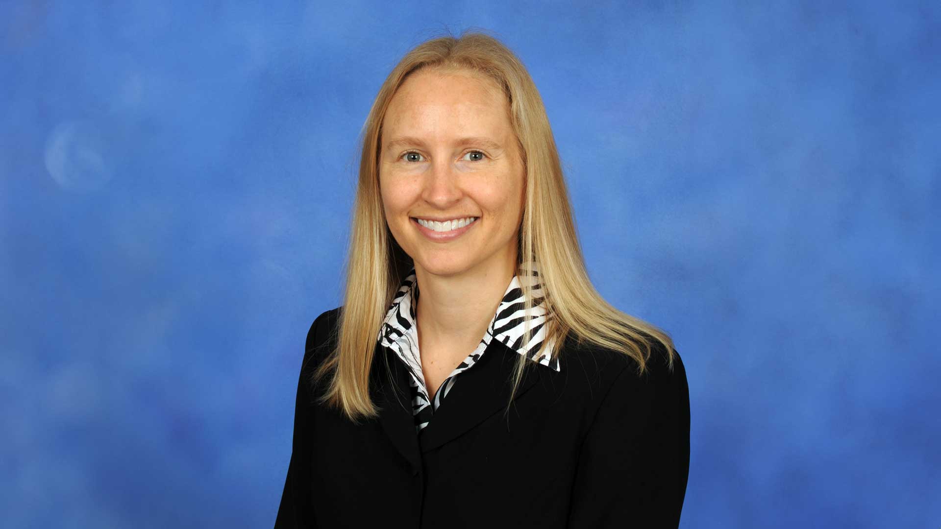 A blonde woman with a black jacket poses in for a headshot in front of a blue background.