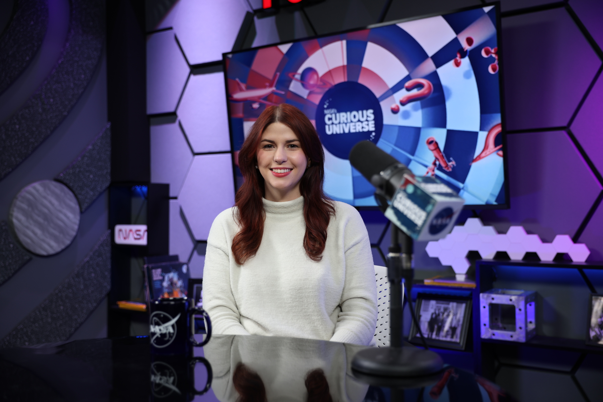 Katie Konans sits at a dark-colored desk wearing a white sweater with long brown hair. Behind her is a TV screen with the Curious Universe logo.