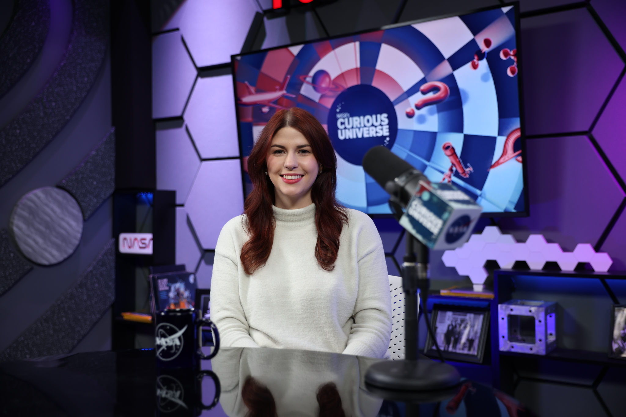 Katie Konans sits at a dark-colored desk wearing a white sweater with long brown hair. Behind her is a TV screen with the Curious Universe logo.