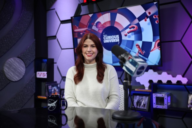 Katie Konans sits at a dark-colored desk wearing a white sweater with long brown hair. Behind her is a TV screen with the Curious Universe logo.