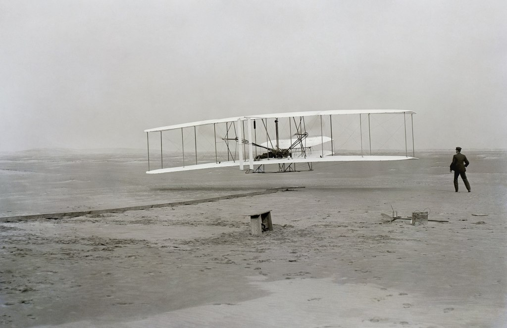 Orville Wright during the first powered flight of a heavier-than-air aircraft; Wilbur is standing to the right of the aircraft