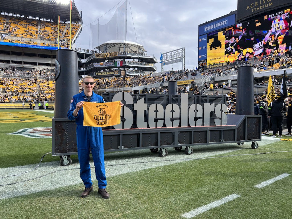 Astronaut Woody Hoburg, wearing a blue NASA flight suit and sunglasses, holds a yellow ceremonial towel at Acrisure Stadium in Pittsburgh. He is posing on the field in front of a large “Steelers” sign and stands filled with fans.