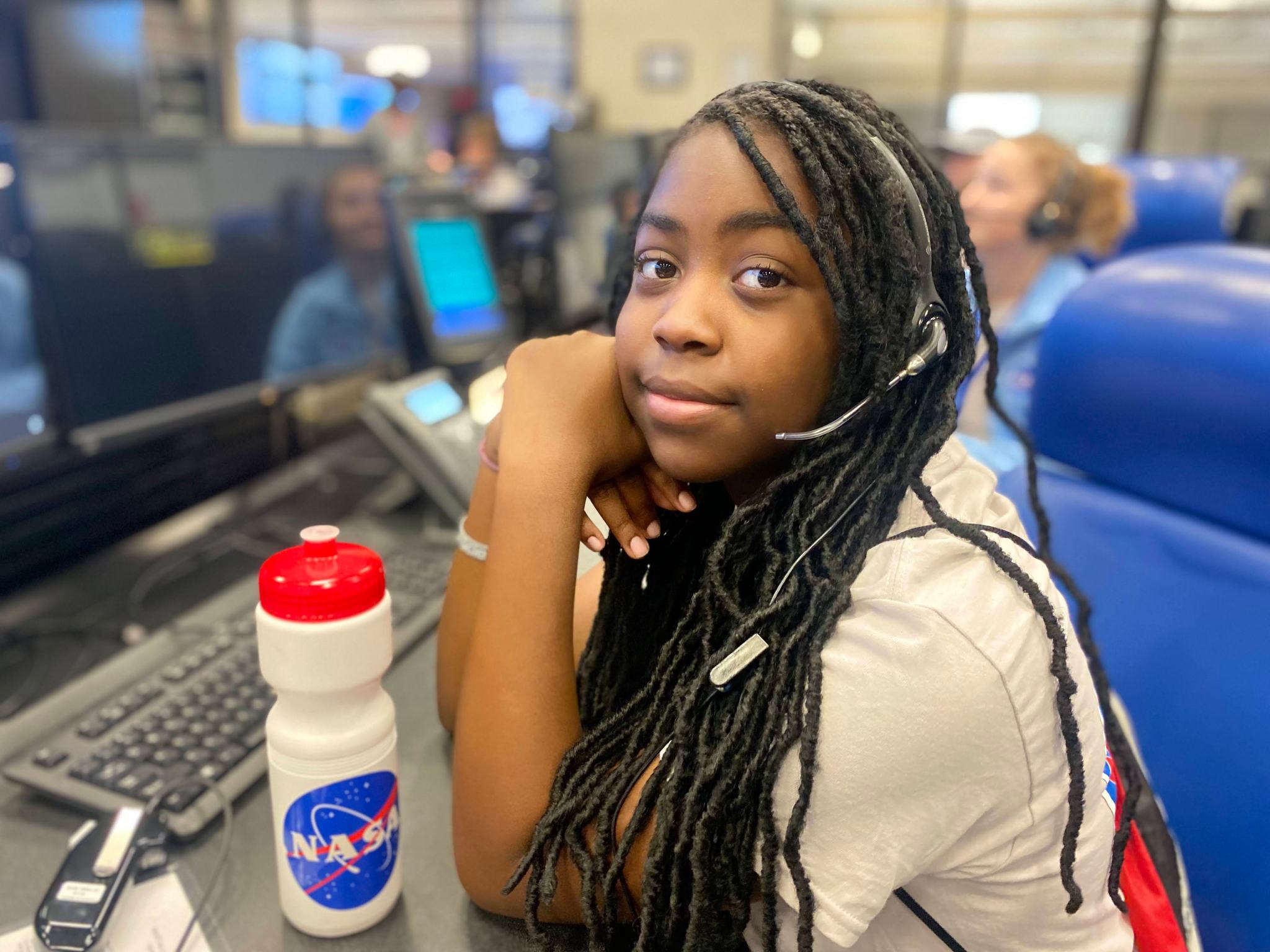 A girl wearing a headset looks towards the camera while sitting in front a a computer console in the range control center. A white water bottle with a blue, red and white NASA meatball sticker sits on the desk in front of her. The red top of the water bottle is closed tightly.