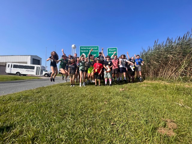a group of 30 students jump into the air in front of two green and white signs that read "International Space Station On-Ramp" and "Moon Ahead"