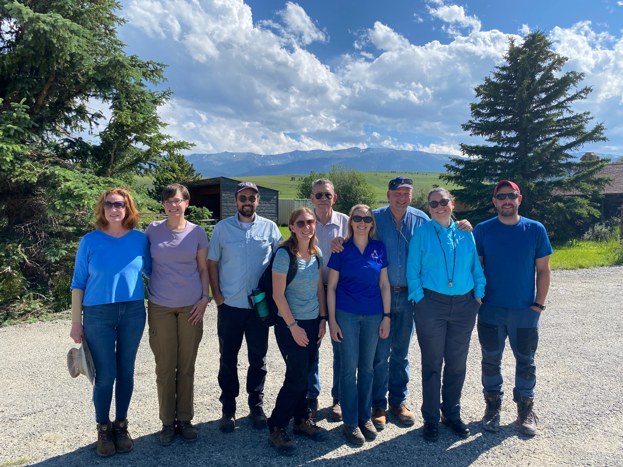 A group of people, some wearing sunglasses, all wearing blue shirts stand on a gravel lot outside with a blue sky and green trees behind them.