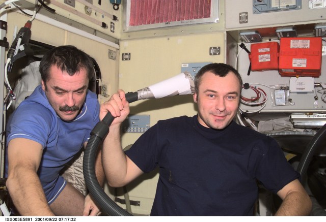Cosmonauts Mikhail Tyurin (left) and Vladimir N. Dezhurov, Expedition Three flight engineers representing Rosaviakosmos, take turns cutting each other’s hair in the Zvezda Service Module on the International Space Station (ISS). Dezhurov holds a vacuum device the crew has fashioned to garner freshly cut hair floating freely. This image was taken with a digital still camera.