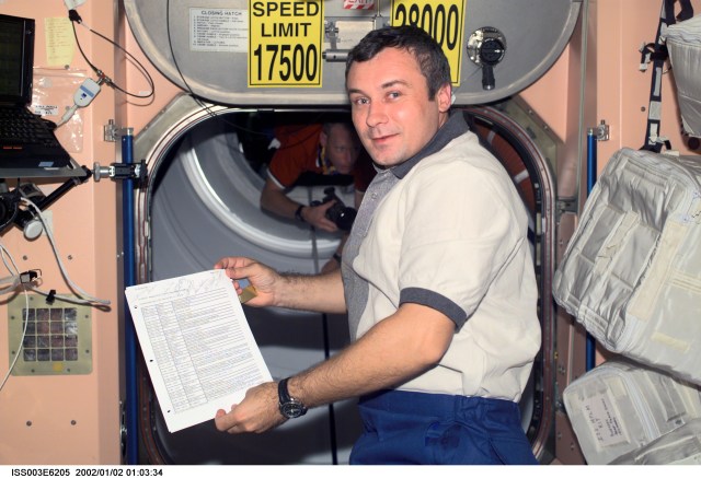 Cosmonaut Vladimir N. Dezhurov, Expedition Three flight engineer representing Rosaviakosmos, holds a document in the Unity node on the International Space Station (ISS). Astronaut Patrick G. Forrester, STS-105 mission specialist, is partly visible in the background. This image was taken with a digital still camera.