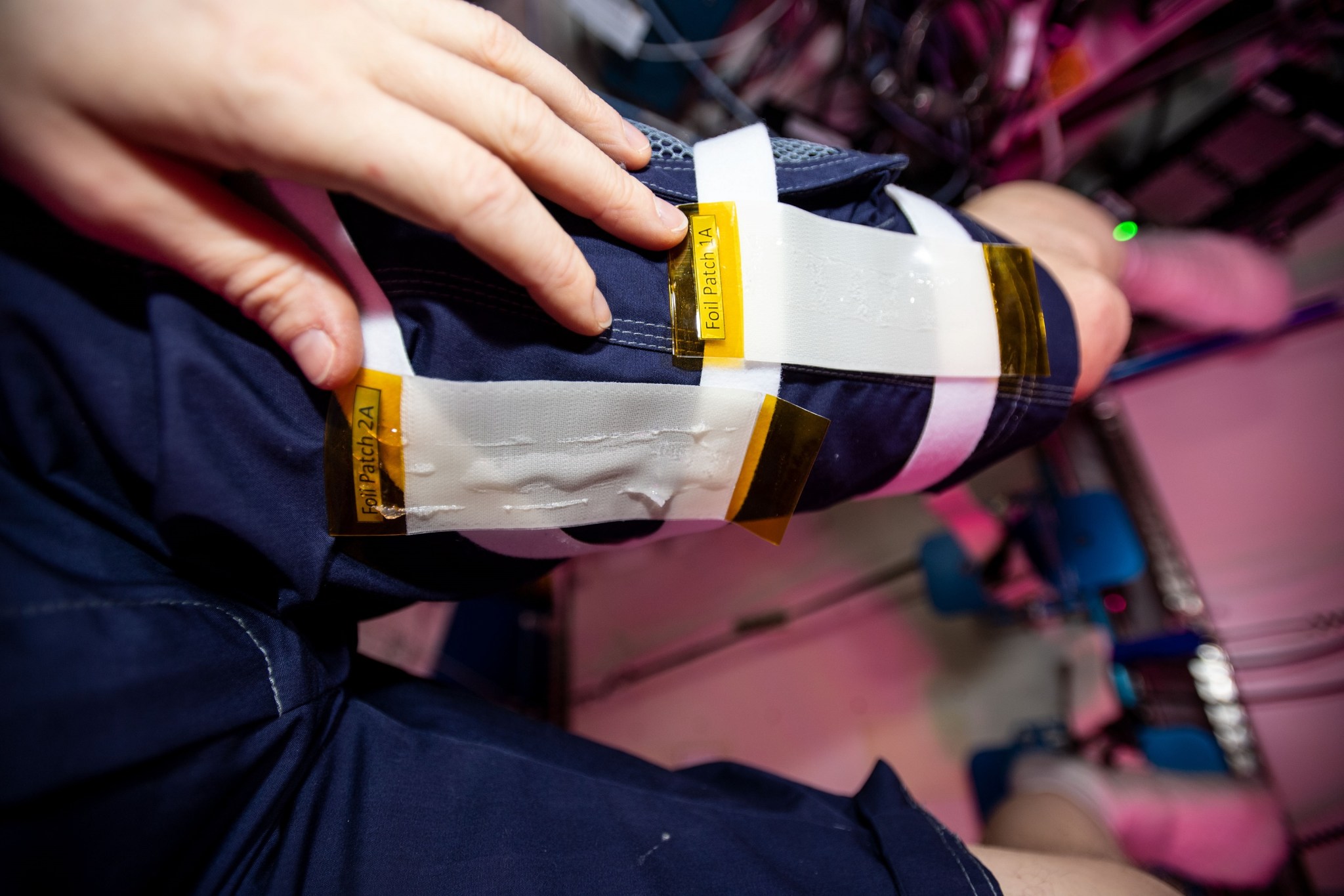 Two white patches the size of a small envelope, labeled “Foil Patch 1A” and “Foil Patch 2A,” are stuck to an astronaut’s blue pants just above the knee. The astronaut’s hand is resting next to the patches.