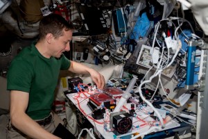 Hoburg adjusts part of an experiment on the table in front of him. He is wearing a green t-shirt and khakis. Two transparent containers of red liquid are attached to transparent tubes running in different directions.