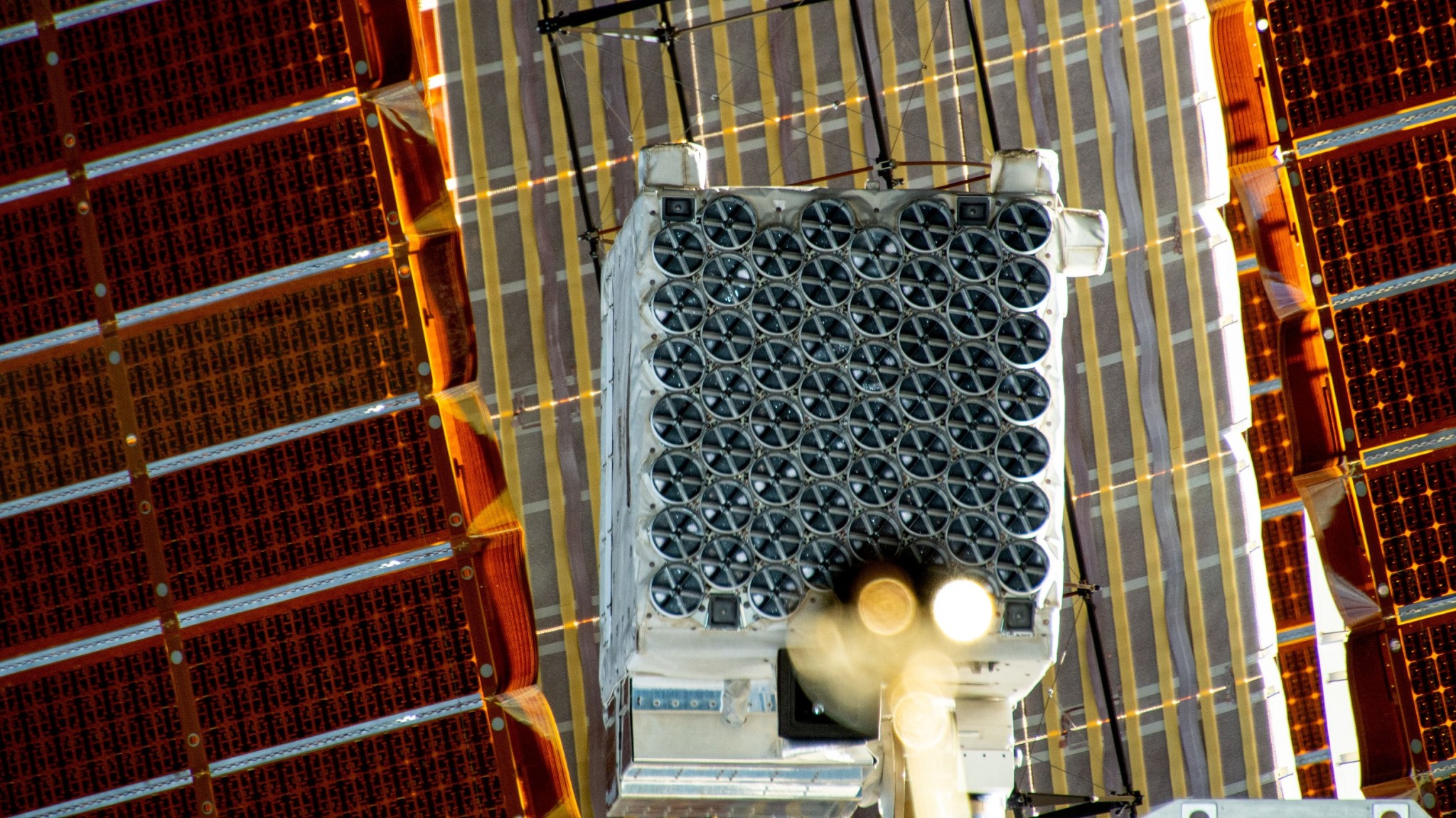 Una gran caja blanca unida al exterior de la estación espacial está cubierta con detectores circulares que parecen ruedas pequeñas. Los paneles solares de la estación ocupan el fondo.