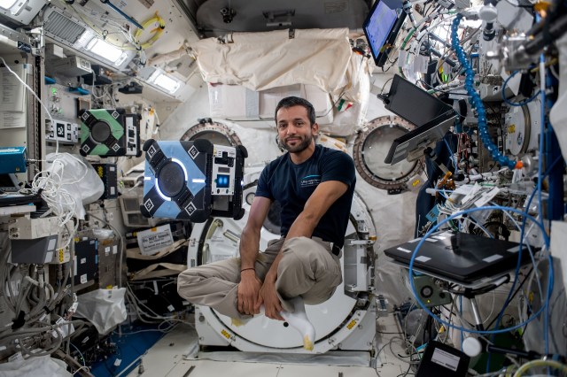 Alneyadi, wearing a dark blue shirt, khaki pants, and white socks, floats with his legs crossed to the right of the blue cube-shaped Astrobee robot. A green Astrobee floats in the background. Laptops, cords, lights, and equipment cover the walls around him.