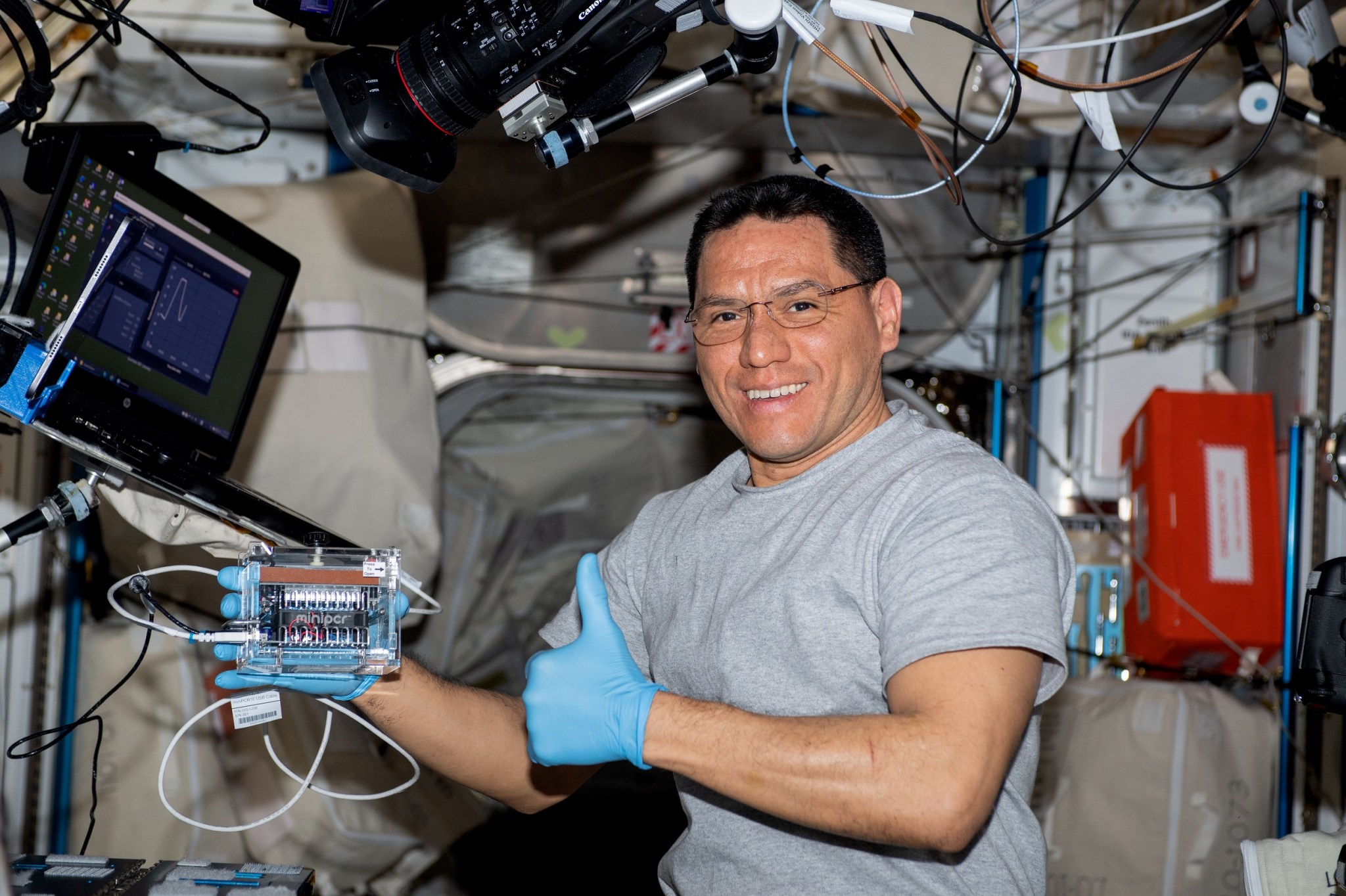 Rubio tiene guantes azules de goma y levanta el pulgar mientras sostiene una pequeña máquina para pruebas de PCR del tamaño de un libro de bolsillo dentro del módulo del laboratorio Columbus. A su derecha está una computadora portátil y sobre él hay una cámara.