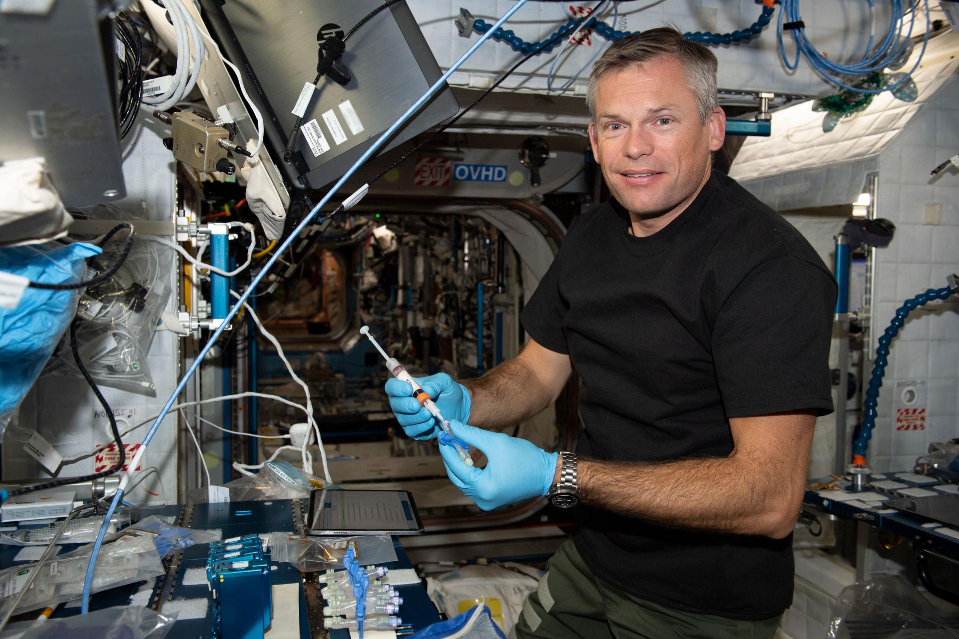 : ESA astronaut Andreas Mogensen, wearing a black t-shirt and green pants, holds a syringe and smiles at the camera. He is holding a syringe with both gloved hands. Several vials are taped to the workbench in front of him.