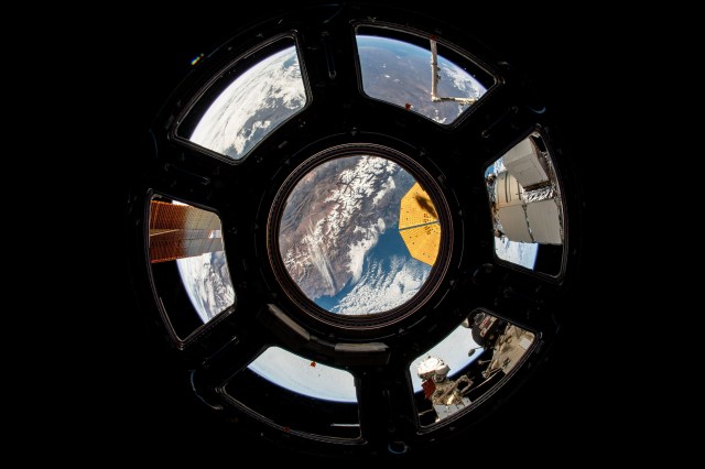 The western coast of Chile is visible through the middle window of the cupola. A solar array can be seen through a window on the left and in the center, a portion of Northrop Grumman's cymbal-shaped solar array. The Roscosmos segment of the station is visible in the bottom right.