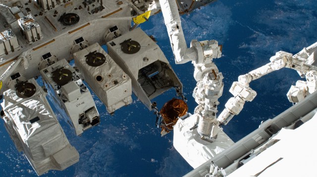 The long white Canadarm2 and Japanese robotic arm manipulate a large white package into a new terminal on the outside of the space station. The blue Earth is visible below with scattered thin clouds.