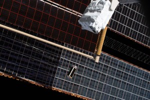 A CubeSat, a rectangular box with black panels on each side, just released from the white satellite deployer at the top right of the image, moves in front of the station’s large solar panels, with the blackness of space visible behind them,