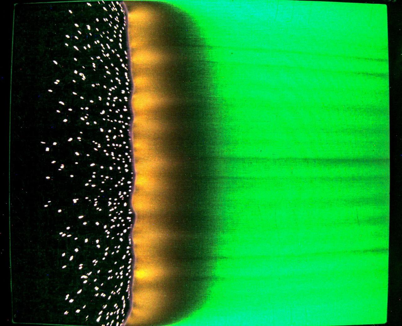 A sample of fabric burns inside Spacecraft Fire Experiment-IV (Saffire-IV). The sample is a composite fabric made of cotton and fiberglass and is 40 cm wide. The image appears green on the right because green LED lights are used to illuminate the sample during the burn. An orange flame sits top to bottom in the center of the image with a dark region between the orange and green areas. Bright specks on a black background to the left of the orange area show the smoldering cotton that remains on the fiberglass substrate after the flame passes