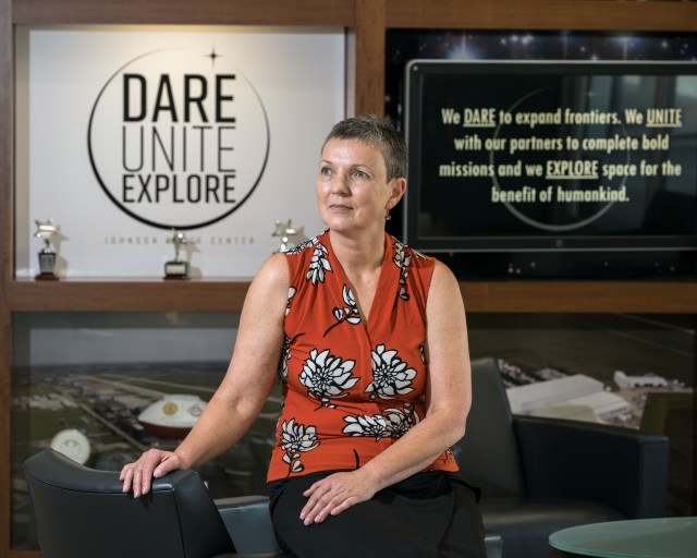 Margarita Sampson thoughtfully gazes off into the distance as she sits in the lobby of Building 1 at NASA's Johnson Space Center. Behind her are two pieces of text. The first says, "Dare Unite Explore" in a circle above "Johnson Space Center." The second says, "We DARE to expand frontiers. We UNITE with our partners to complete bold missions and we EXPLORE space for the benefit of humankind."