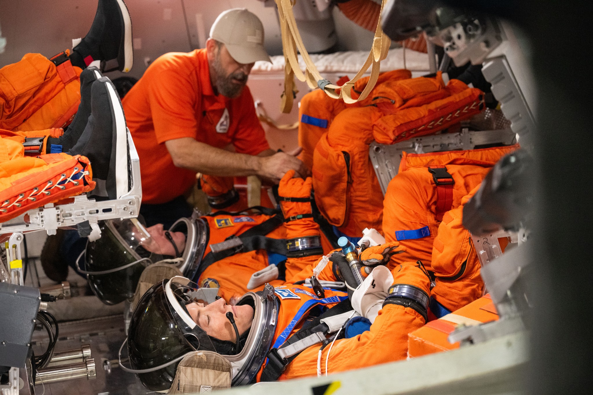 Two astronauts in orange spacesuits and helmets sit horizontal with their feet raised in a mockup of the Orion spacecraft. The feet of two other astronauts are above them on the left. All are assisted by a man in a beige hat.
