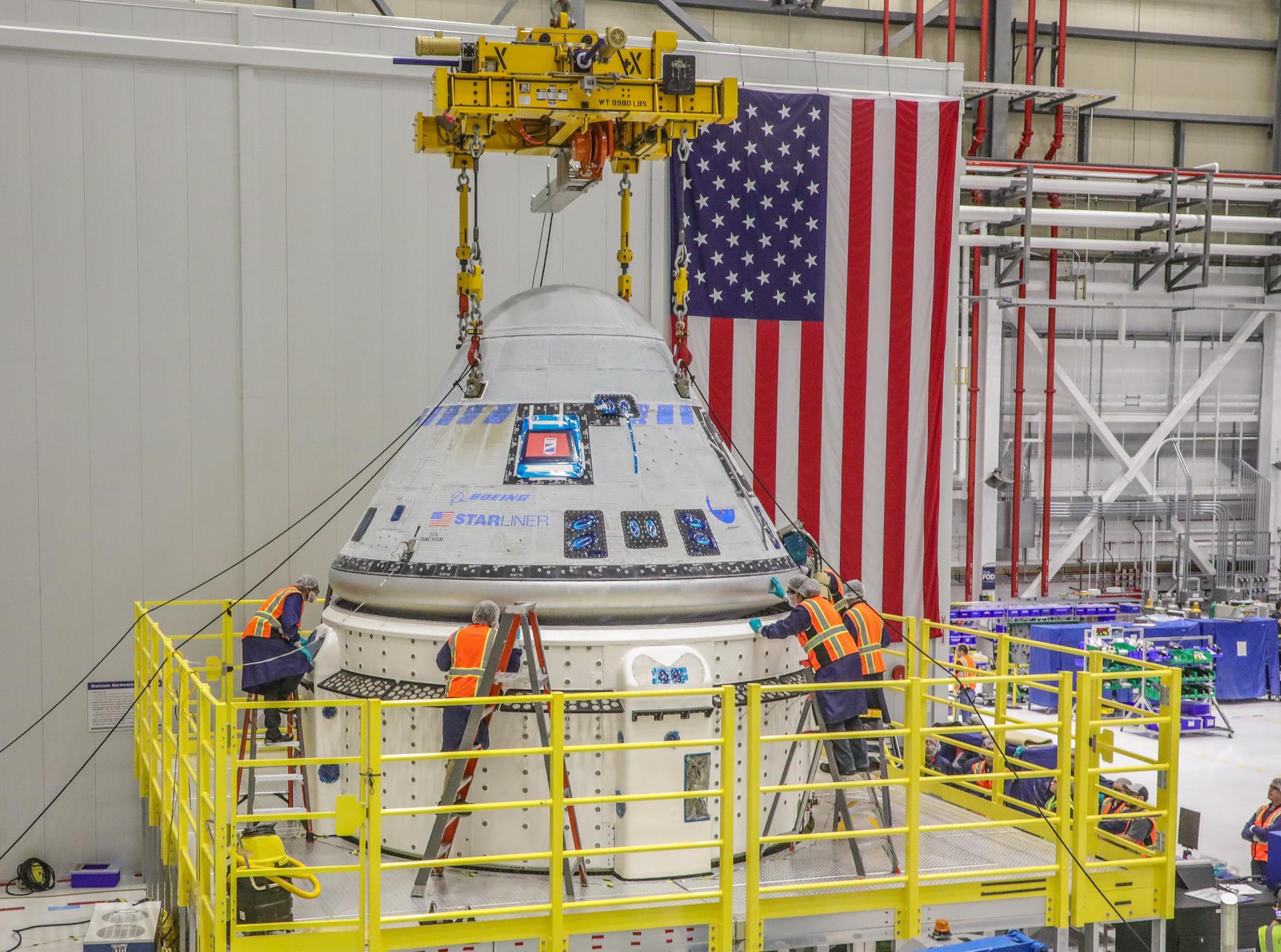 Technicians work around Boeing's Starliner spacecraft supported by a yellow crane during processing.