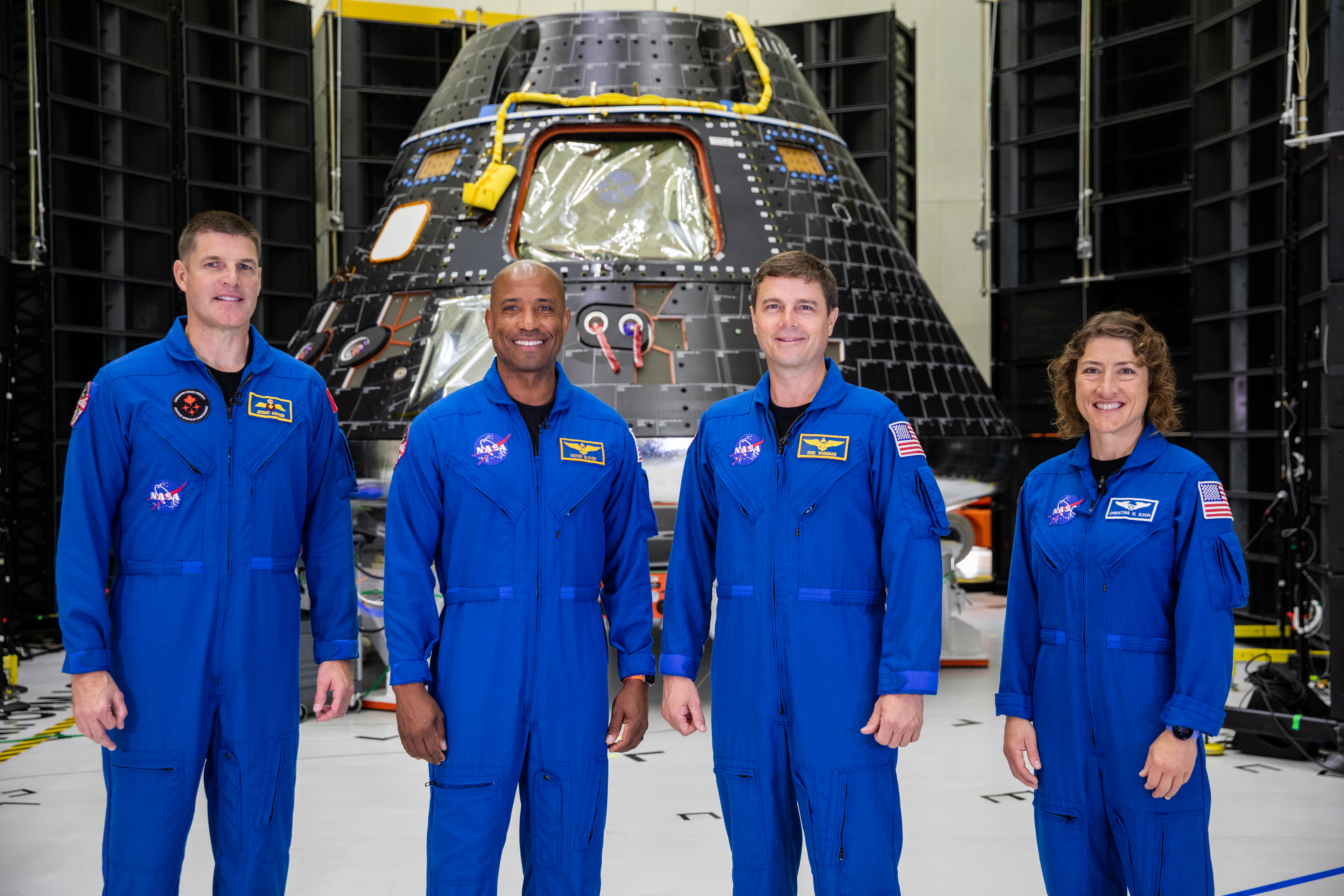 Four astronauts wearing blue flight suits stand inside a building, with the Orion crew module behind them. They are, from left, Jeremy Hansen, Victor Glover, Reid Wiseman, and Christina Koch. Glover is a Black man, and Koch is a White woman. Orion is cone-shaped, with a flat top. It is black and has yellow wiring on it, as well as blue dots around some panels.