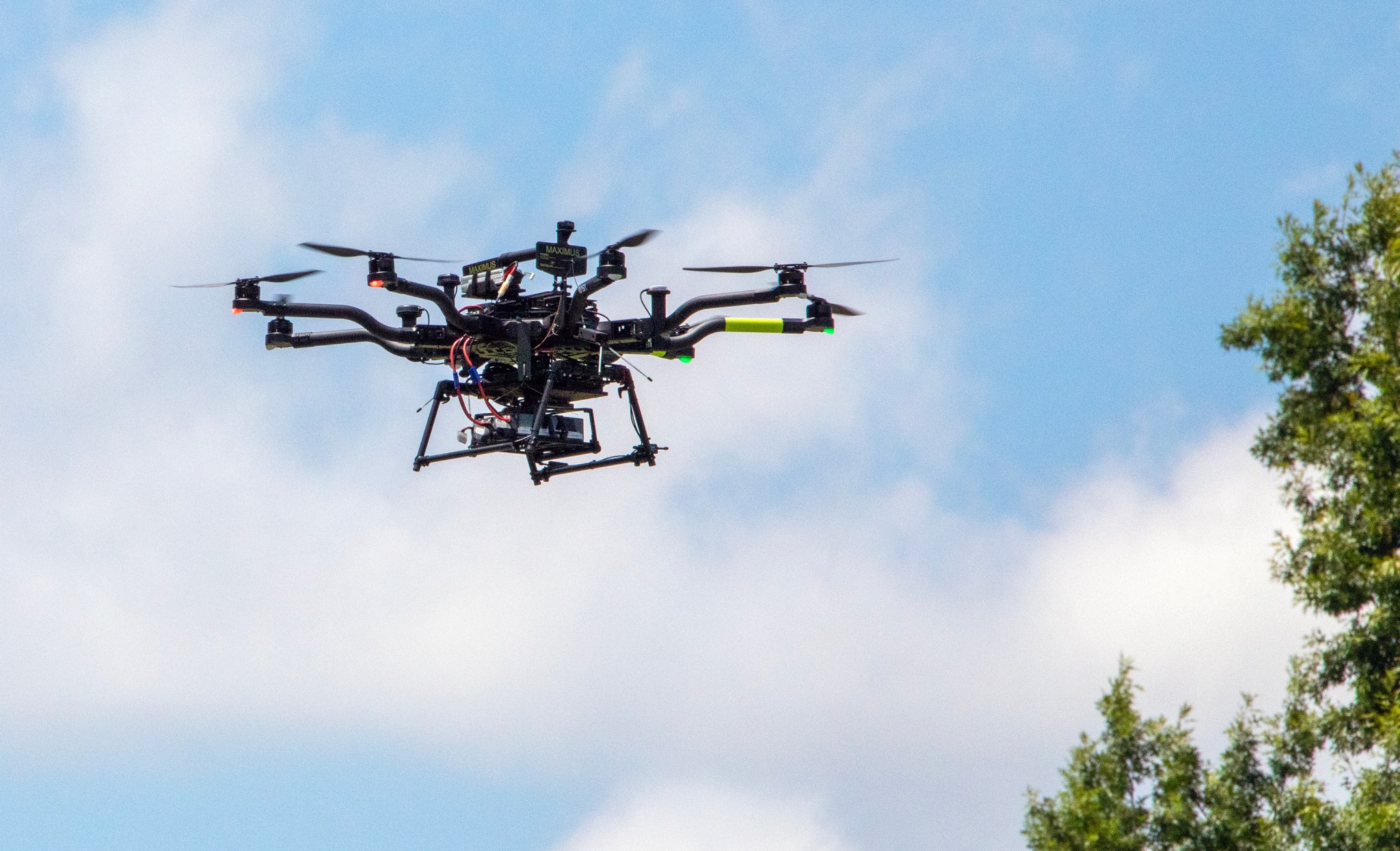 A small, black drone with multiple helicopter-like blades hovers over some trees during a bright, partly cloudy day in Virginia.