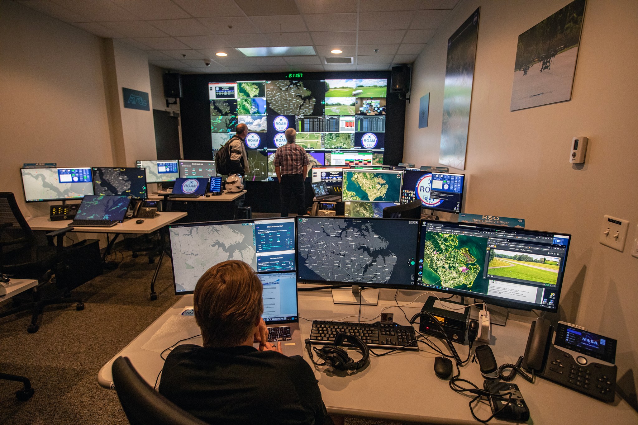 Uma sala cheia de telas de computador sobre mesas e uma parede oposta são vigiadas por pesquisadores que monitoram o voo de pequenos drones.