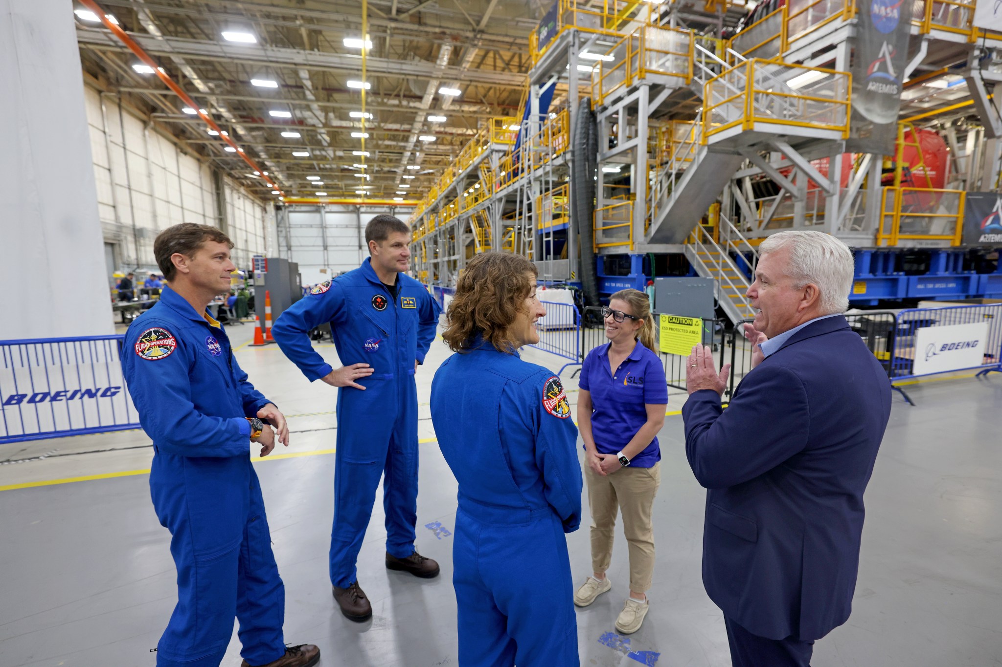 From left to right, NASA astronaut Reid Wiseman, CSA astronaut Jeremy Hansen, NASA astronaut Christina Koch, and two people stand in a circle as they have a discussion. The astronauts wear blue flight suits with various patches on them. A building with bare ceilings and concrete floors stretches far behind them.