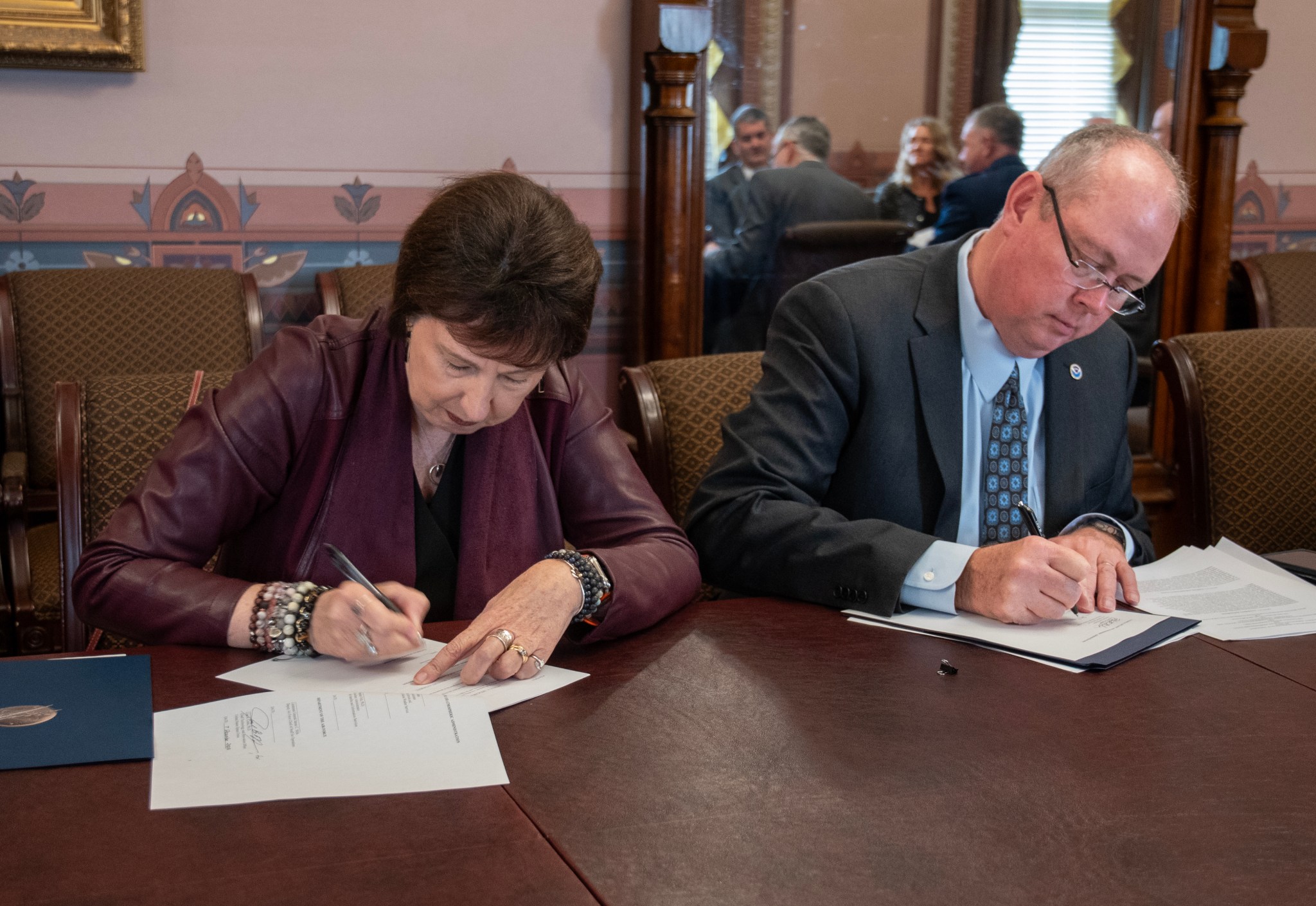 Two people sit at a brown table. Nicola Fox is sitting on the left, signing a sheet of paper. On the right, Ken Graham is doing the same.