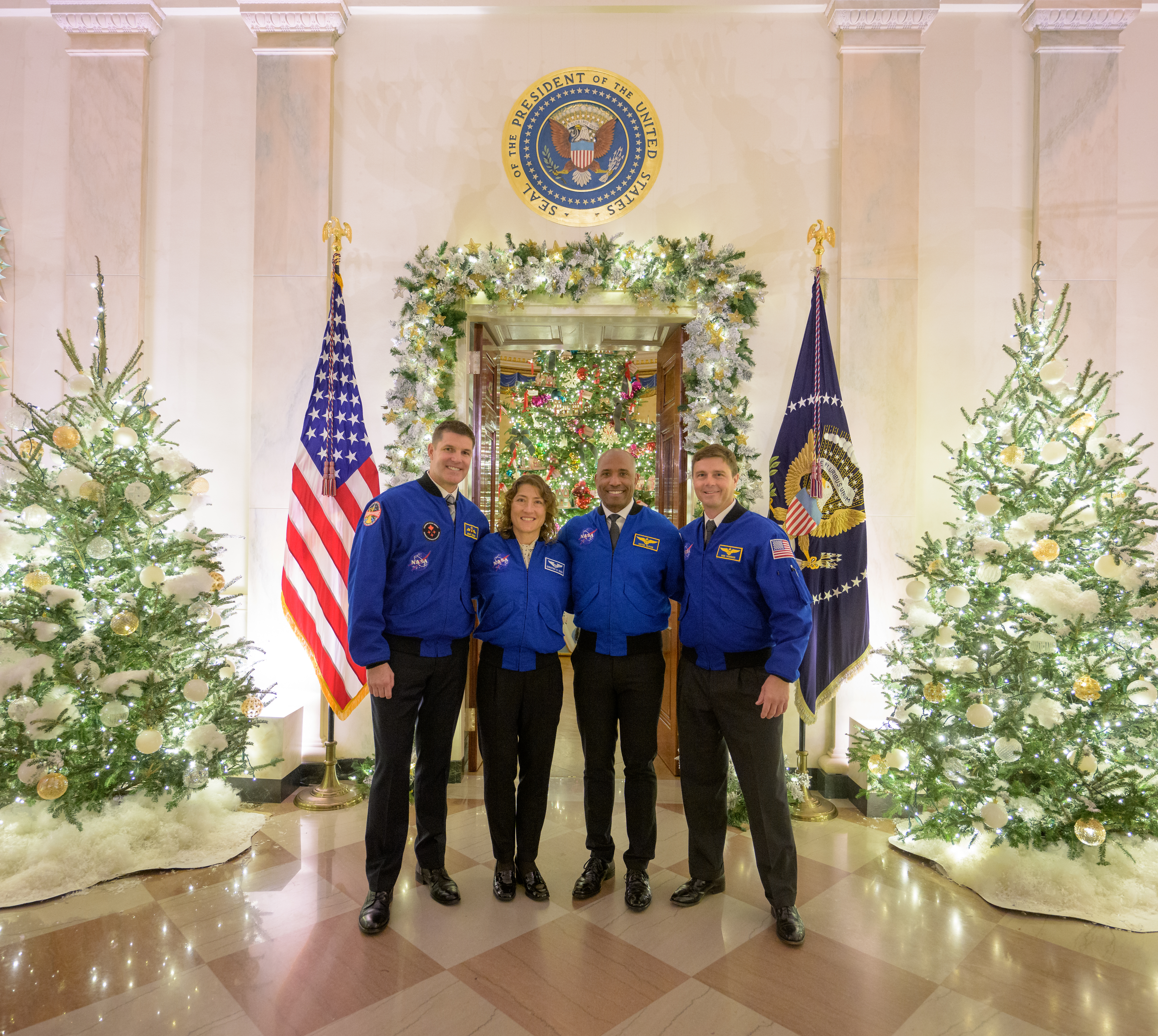 Four astronauts, wearing blue jackets and black pants, pose for a photo at the White House in front of decorations like trees, wreaths, and boughs. The astronauts, members of the Artemis II mission, have their arms around each other's shoulders. Behind them are two flags, and a seal high on the wall that reads "Seal of the President of the United States."