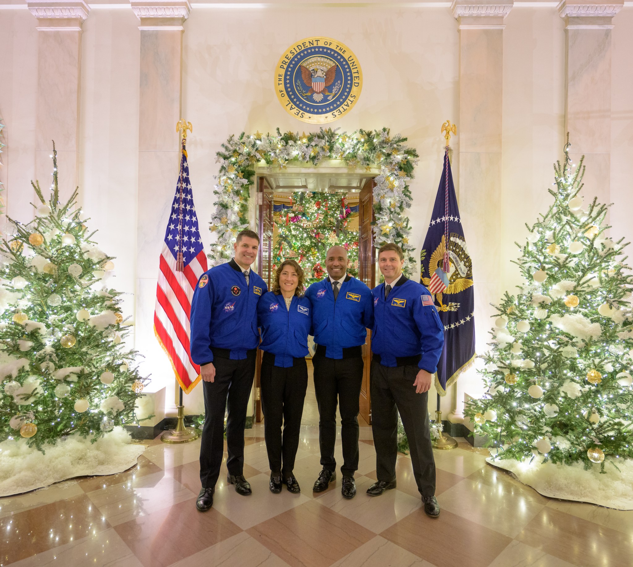 Four astronauts, wearing blue jackets and black pants, pose for a photo at the White House in front of decorations like trees, wreaths, and boughs. The astronauts, members of the Artemis II mission, have their arms around each other's shoulders. Behind them are two flags, and a seal high on the wall that reads 