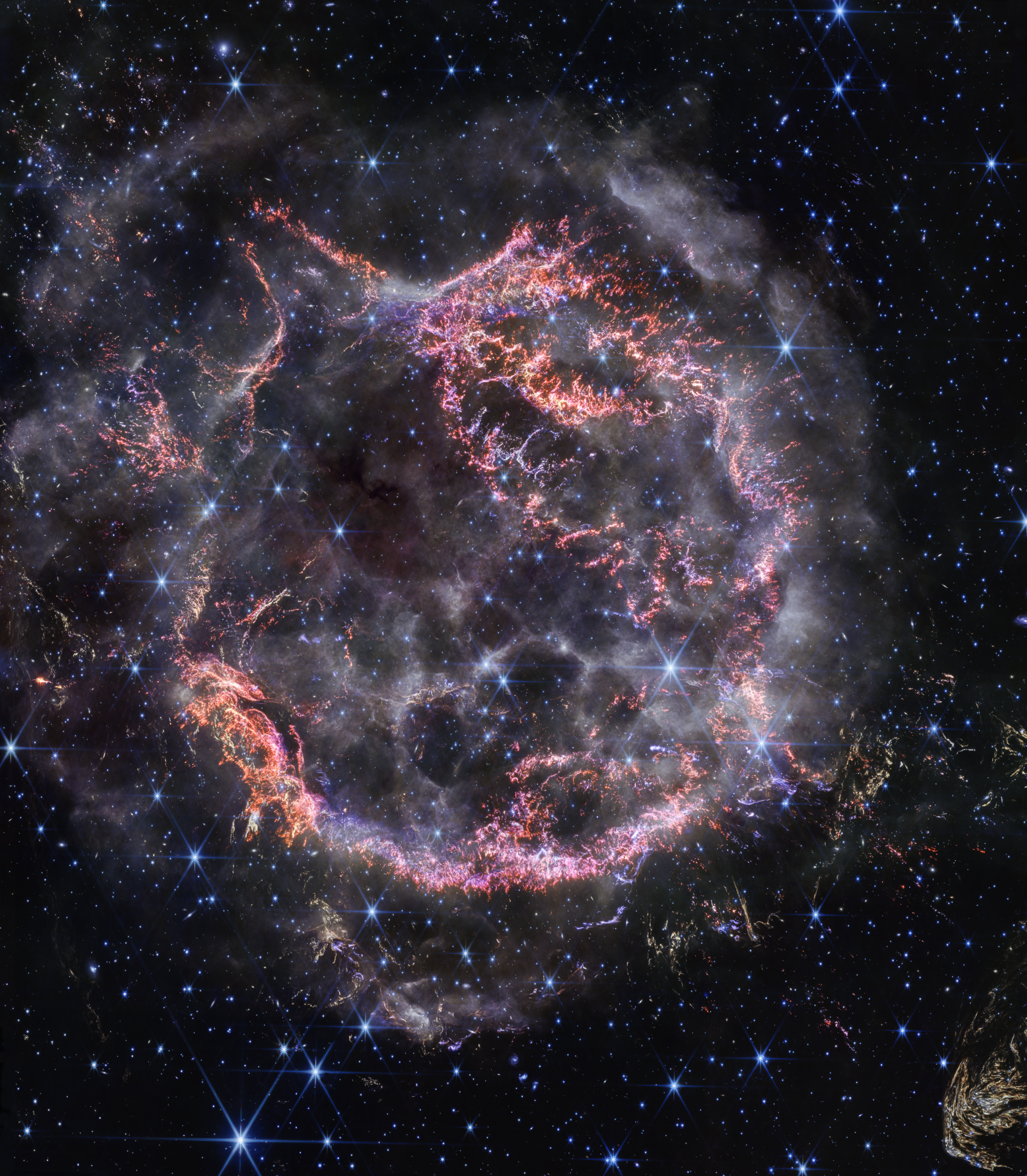 NASA’s James Webb Space Telescope’s new view of Cassiopeia A in near-infrared light is giving astronomers hints at the dynamical processes occurring within the supernova remnant. Tiny clumps represented in bright pink and orange make up the supernova’s inner shell, and are comprised of sulfur, oxygen, argon, and neon from the star itself. A large, striated blob at the bottom right corner of the image, nicknamed Baby Cas A, is one of the few light echoes visible NIRCam’s field of view. In this image, red, green, and blue were assigned to Webb’s NIRCam data at 4.4, 3.56, and 1.62 microns (F444W, F356W, and F162M, respectively).