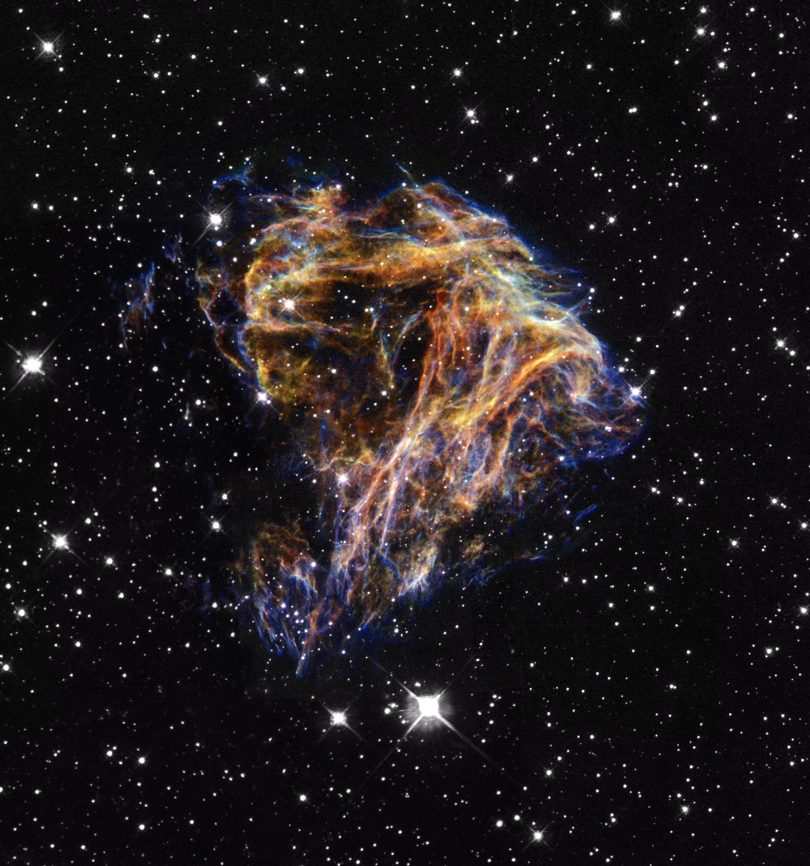 Many points of light are scattered across the black background of space like confetti. At center are blue and orange strands, debris from a star explosion.
