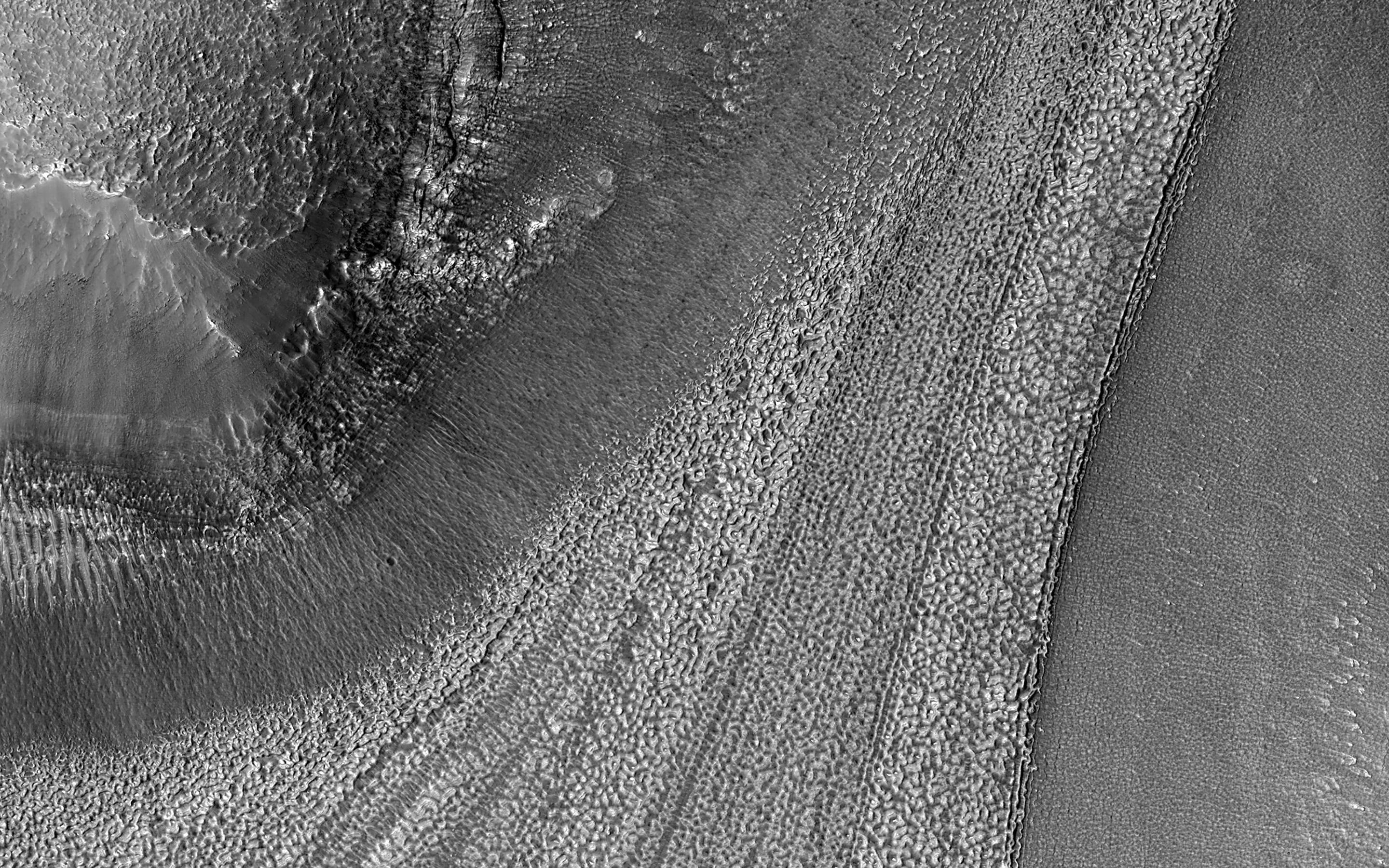 A black and white image of a portion of Mars' surface from above. Ridges stretch from top right to the bottom, the result of ice moving over possibly thousands of years. The glacier-like forms left behind have a mottled appearance.