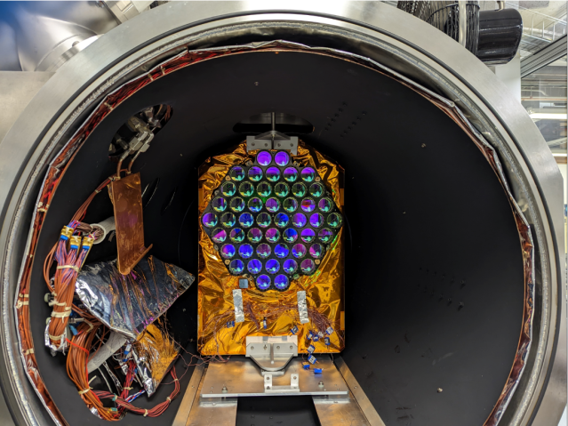 The laser retroreflector array is sitting inside a large testing chamber. The array is covered in a copper foil-like material. The mirrors of the array are placed in a honeycomb pattern and are reflecting back purple, blue, and black colors to the camera. These are the colors of what is surrounding the camera.