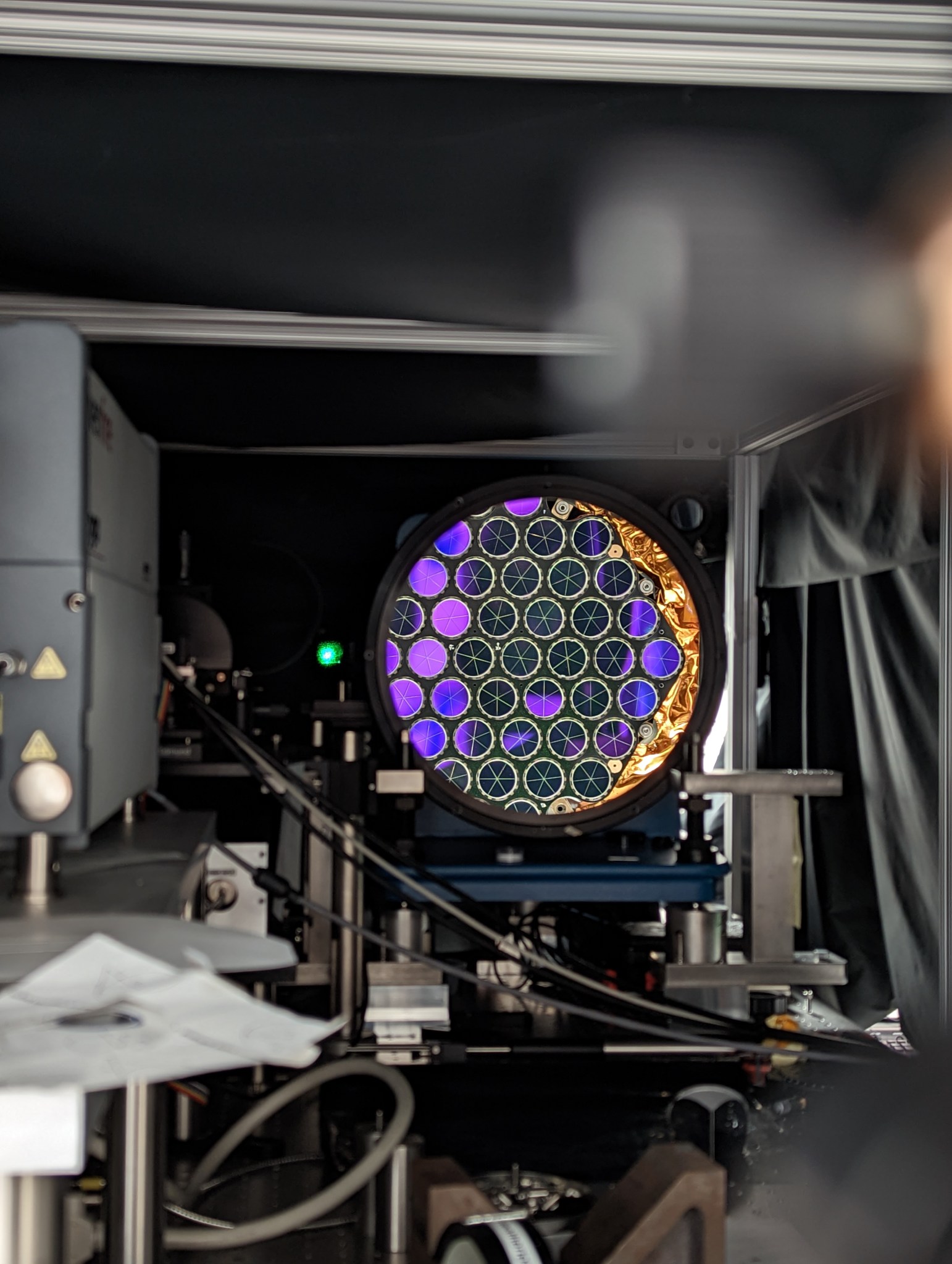 The focus of the image is a circular mirror. The reflection off of the mirror is the laser retroreflector array, which is made up of multiple smaller circular shaped mirrors together in a honeycomb pattern. They are reflecting black and purple colors of the arrays surroundings. At the right edge of the main reflection there is some copper colored foil also being reflected back towards the camera. Surrounding the large mirror is parts of the test apparatus and a darkly lit room.