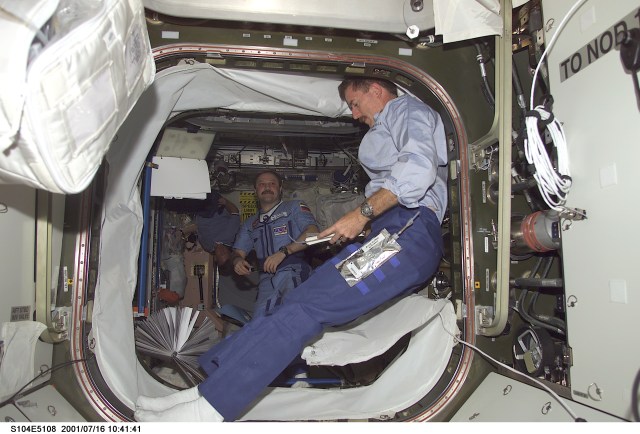 James F. Reilly, STS-104 mission specialist, reads over a checklist in the hatchway of the newly installed Quest Airlock. In the background, cosmonaut Yury V. Usachev of Rosaviakosmos, Expedition Two mission commander, is working in Unity Node 1.