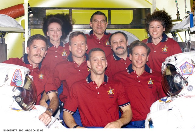 Seven astronauts and a cosmonaut representing Rosaviakosmos take a break in joint activities involving the Expedition Two and STS-104 crews to pose for an in-flight portrait in the newly delivered Quest Airlock on the International Space Station (ISS). Flanked by two extravehicular mobility unit (EMU) space suits, Charles O. Hobaugh is in front. On the second row are, from the left, James F. Reilly, Steven W. Lindsey, Yury V. Usachev and Michael L. Gernhardt. In the rear are Janet L. Kavandi, James S. Voss and Susan J. Helms. Usachev, commander; along with Voss and Helms, both flight engineers, comprise the Expedition Two crew. Lindsey is STS-104 commander, with Hobaugh serving as pilot. Kavandi, STS-104 flight engineer, is joined by Gernhardt and Reilly as mission specialists on the mission. This image was recorded with a digital still camera.