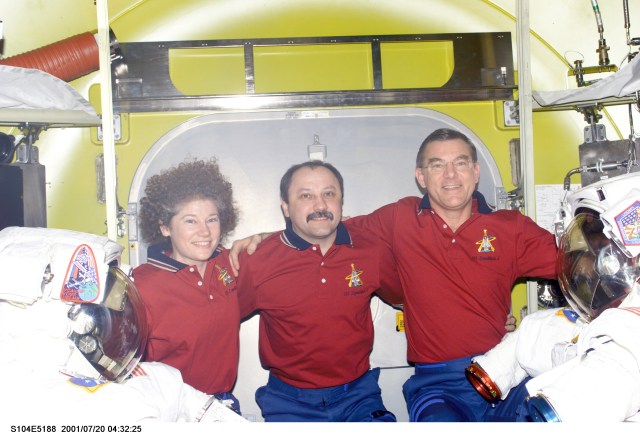 The Expedition Two crew poses for an in-flight portrait in the newly- delivered Quest Airlock on the International Space Station (ISS). Flanked by two extravehicular mobility unit (EMU) space suits, are, from left, Susan J. Helms, Yury V. Usachev and James S. Voss. Usachev is commander and Voss and Helms are both flight engineers. This image was recorded by one of the visiting STS-104 crew members using a digital still camera.