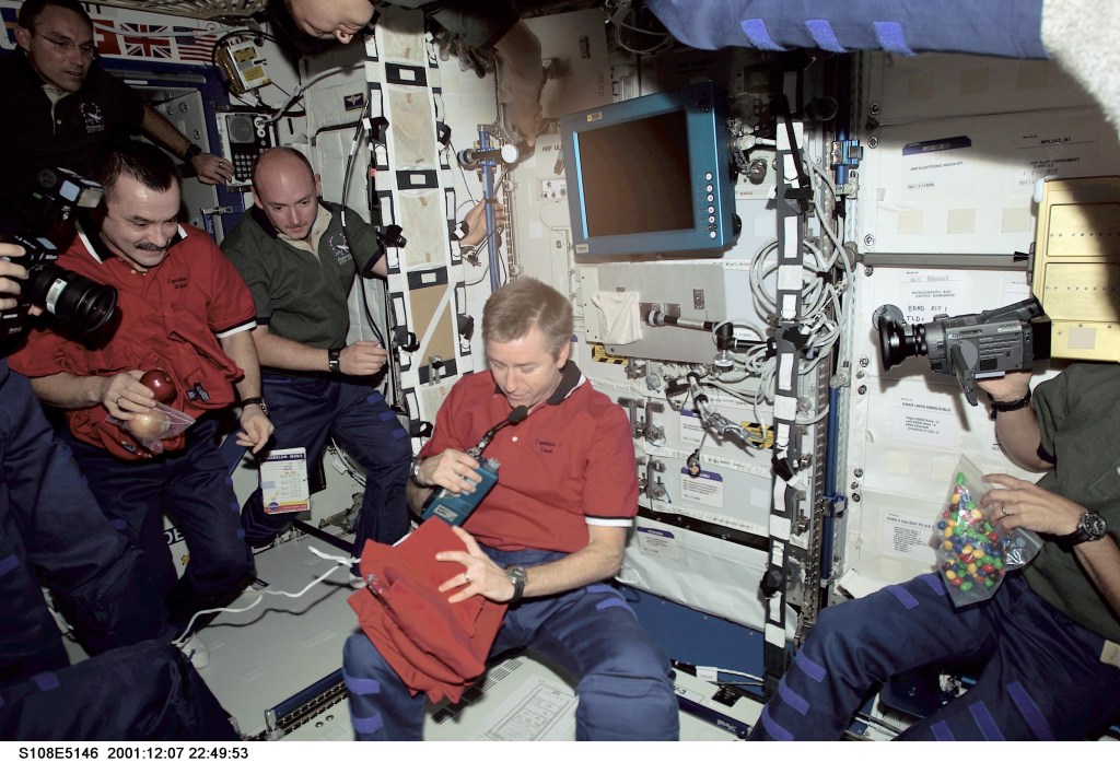 Shortly after the Space Shuttle Endeavour docked with the International Space Station (ISS), astronauts and cosmonauts aboard the two vehicles reunited in the Destiny laboratory. Several unidentified crew members (partially out of frame) aim various kinds of cameras at astronaut Frank L. Culbertson, Jr., Expedition Three commander, as he prepares to continue a station tradition by handing out souvenir shirts from the soon-to-depart station team. Cosmonaut Mikhail Tyurin (left), Expedition Three flight engineer; and astronaut Mark E. Kelly (second left), STS-108 pilot, look on. Hovering above are astronauts Carl E. Walz (in shadowed area, upper left); Expedition Four flight engineer; and Daniel M. Tani, STS-108 mission specialist. The image was taken with a digital still camera.