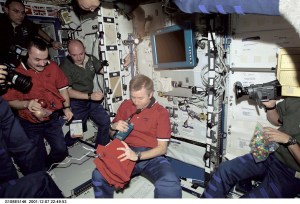 Shortly after the Space Shuttle Endeavour docked with the International Space Station (ISS), astronauts and cosmonauts aboard the two vehicles reunited in the Destiny laboratory. Several unidentified crew members (partially out of frame) aim various kinds of cameras at astronaut Frank L. Culbertson, Jr., Expedition Three commander, as he prepares to continue a station tradition by handing out souvenir shirts from the soon-to-depart station team. Cosmonaut Mikhail Tyurin (left), Expedition Three flight engineer; and astronaut Mark E. Kelly (second left), STS-108 pilot, look on. Hovering above are astronauts Carl E. Walz (in shadowed area, upper left); Expedition Four flight engineer; and Daniel M. Tani, STS-108 mission specialist. The image was taken with a digital still camera.