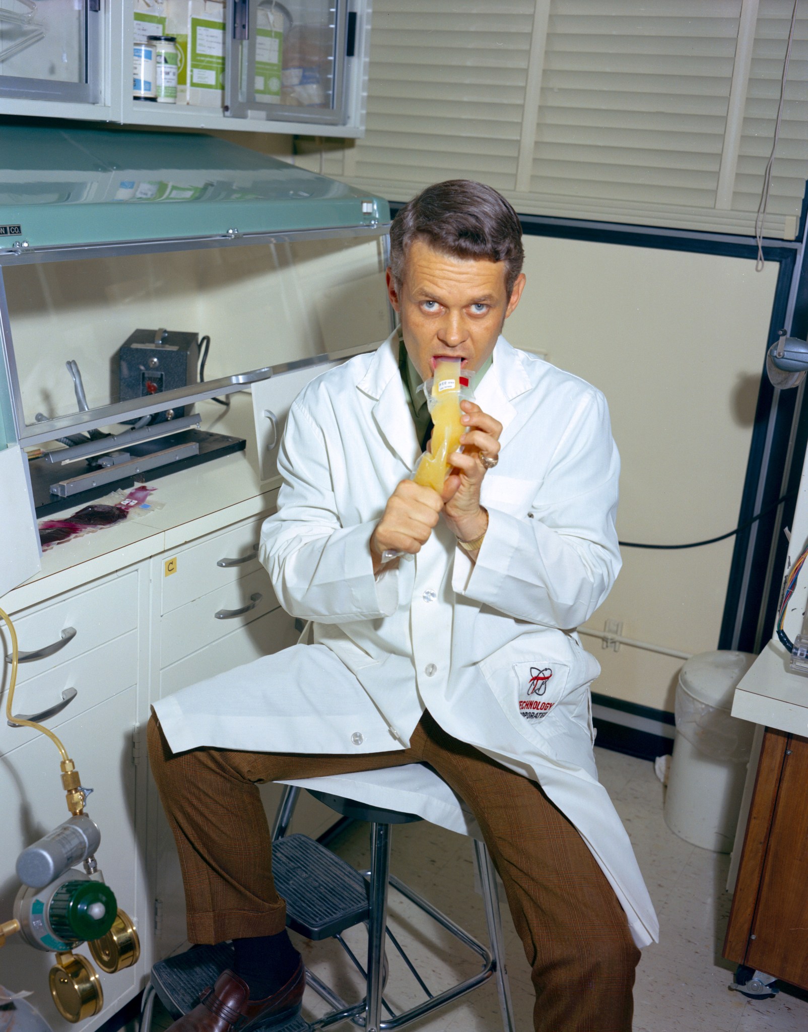 Photo of Malcolm Smith squirting a clear plastic pouch of orange food into his mouth while sitting on a stool.