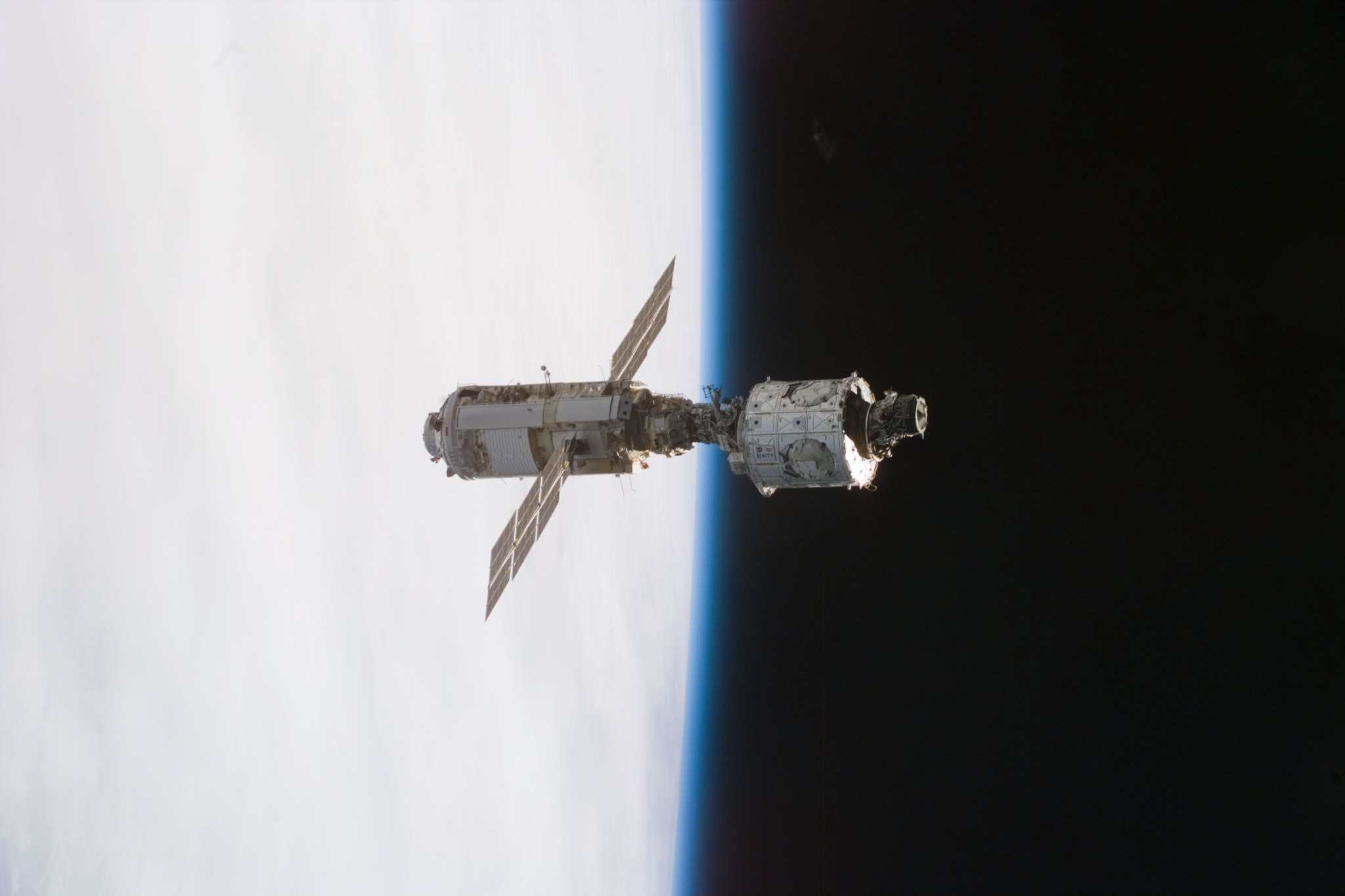 The first pieces of the International Space Station, the mated Russian-built Zarya (left) and U.S.-built Unity modules, are backdropped against the blackness of space and Earth's horizon. Zarya is cylinder-shaped and has solar panels that stick out from the body of the module.