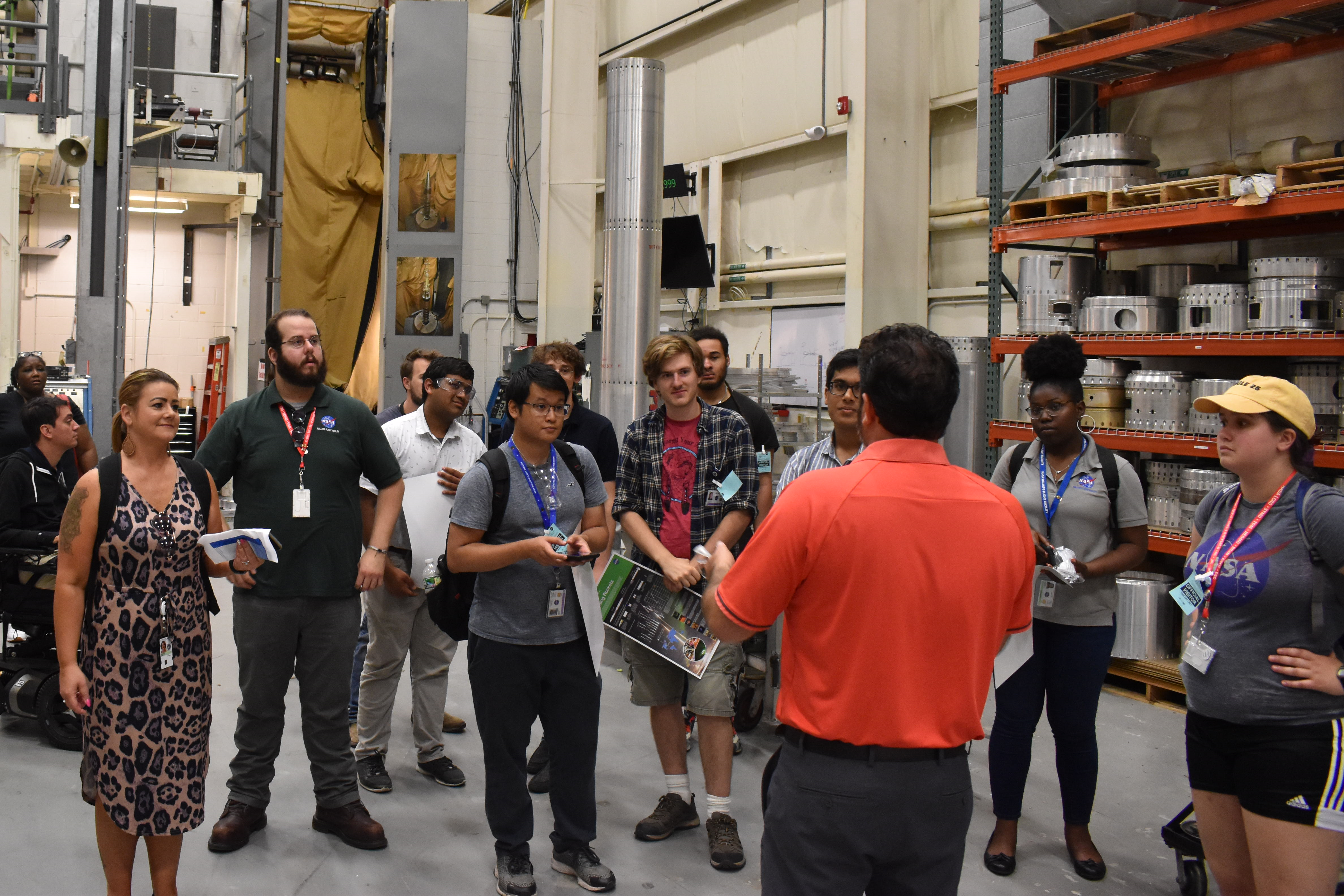 Medium wide perspective of a warehouse area. In the center foreground, a man wearing an orange dress shirt speaks to a group of students.