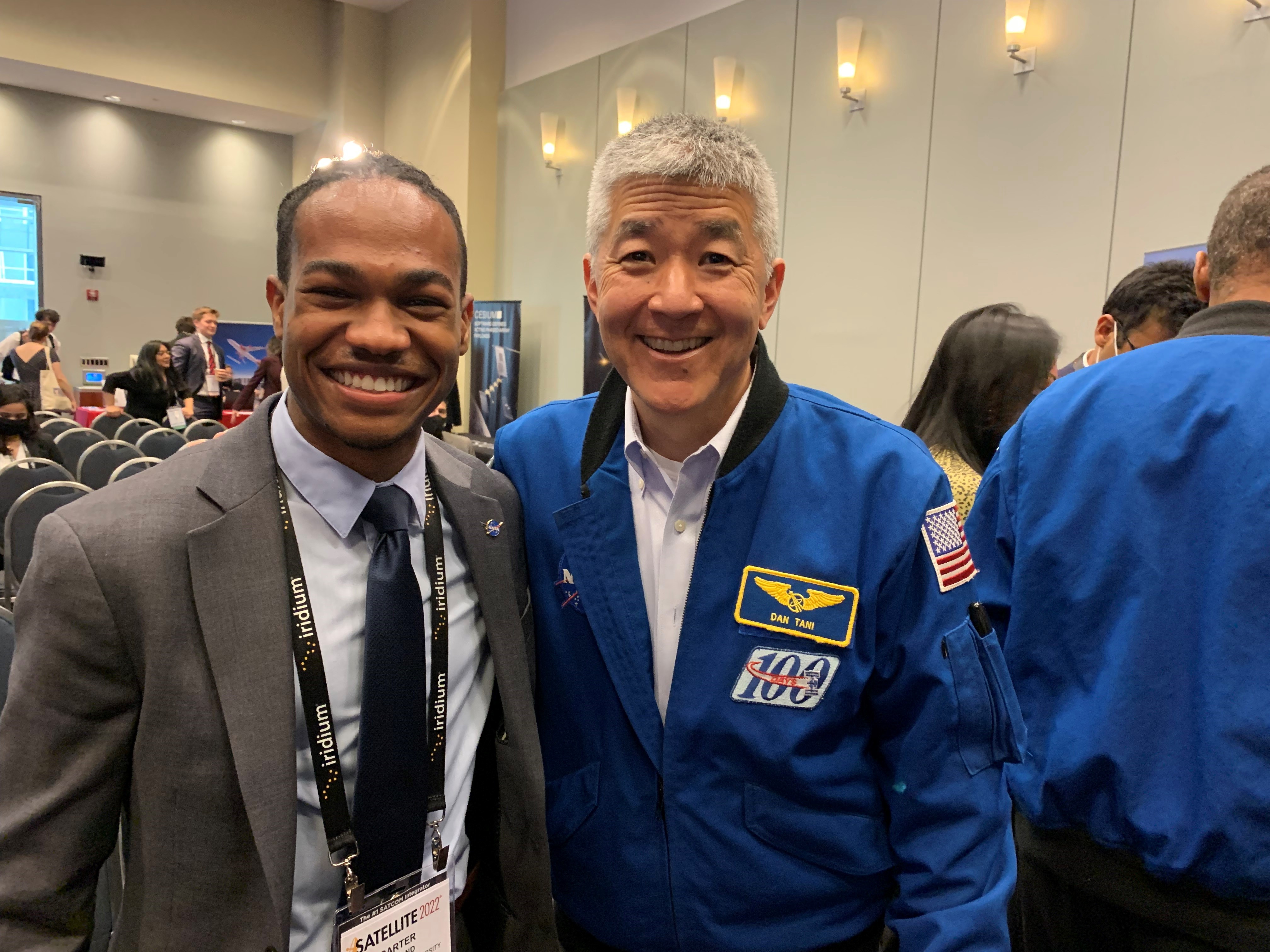 Medium perspective of an auditorium. A student in business attire [left] poses with an astronaut wearing a blue NASA flight suit [right].