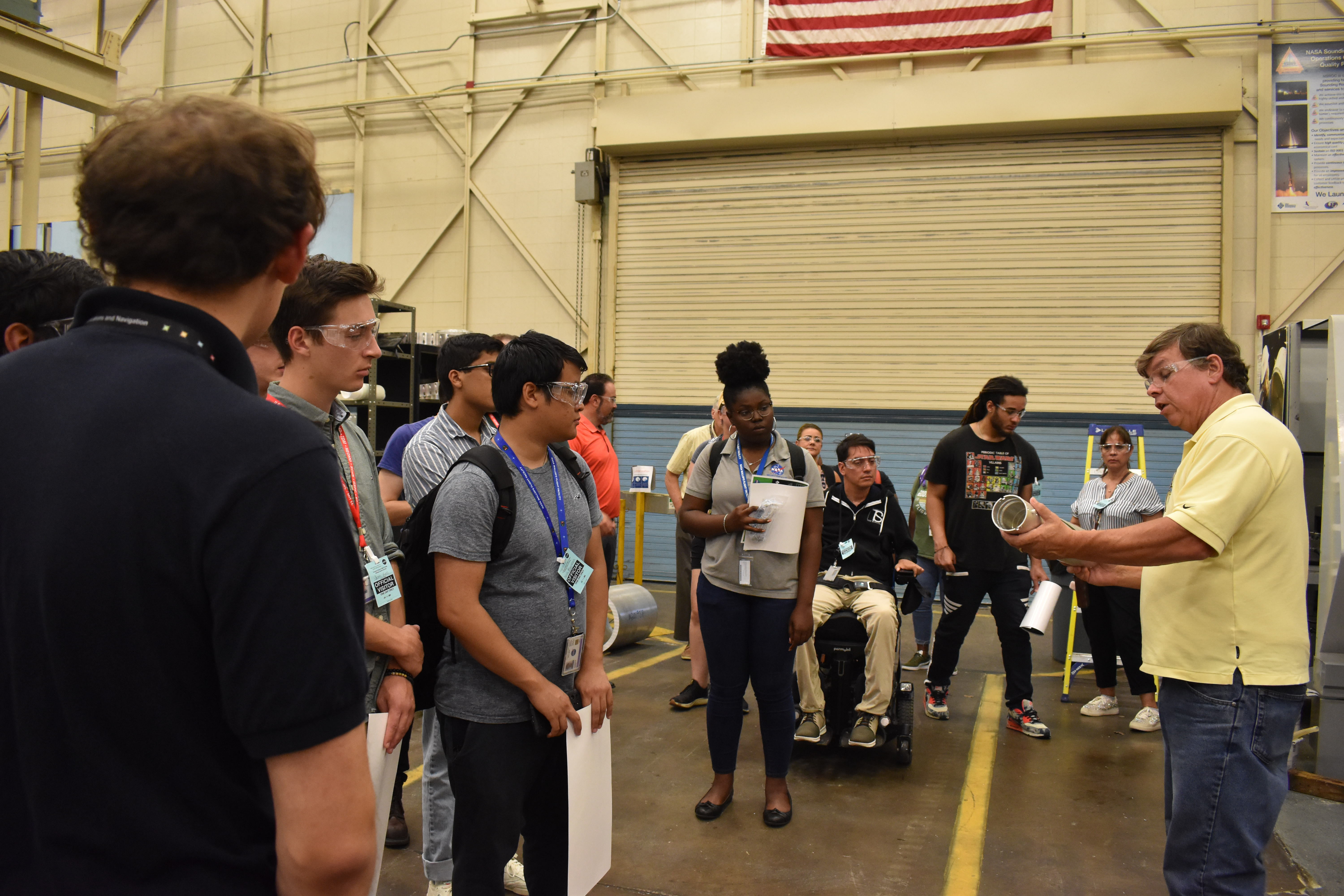 Medium wide perspective of a large warehouse area. In the right foreground, a man wearing goggles and a yellow dress shirt holds a metal cylinder indicating toward it as students look on.