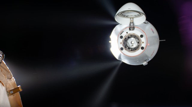 Thrusters on the white SpaceX Dragon appear as four straight white lines coming from behind the spacecraft against the black background of space.