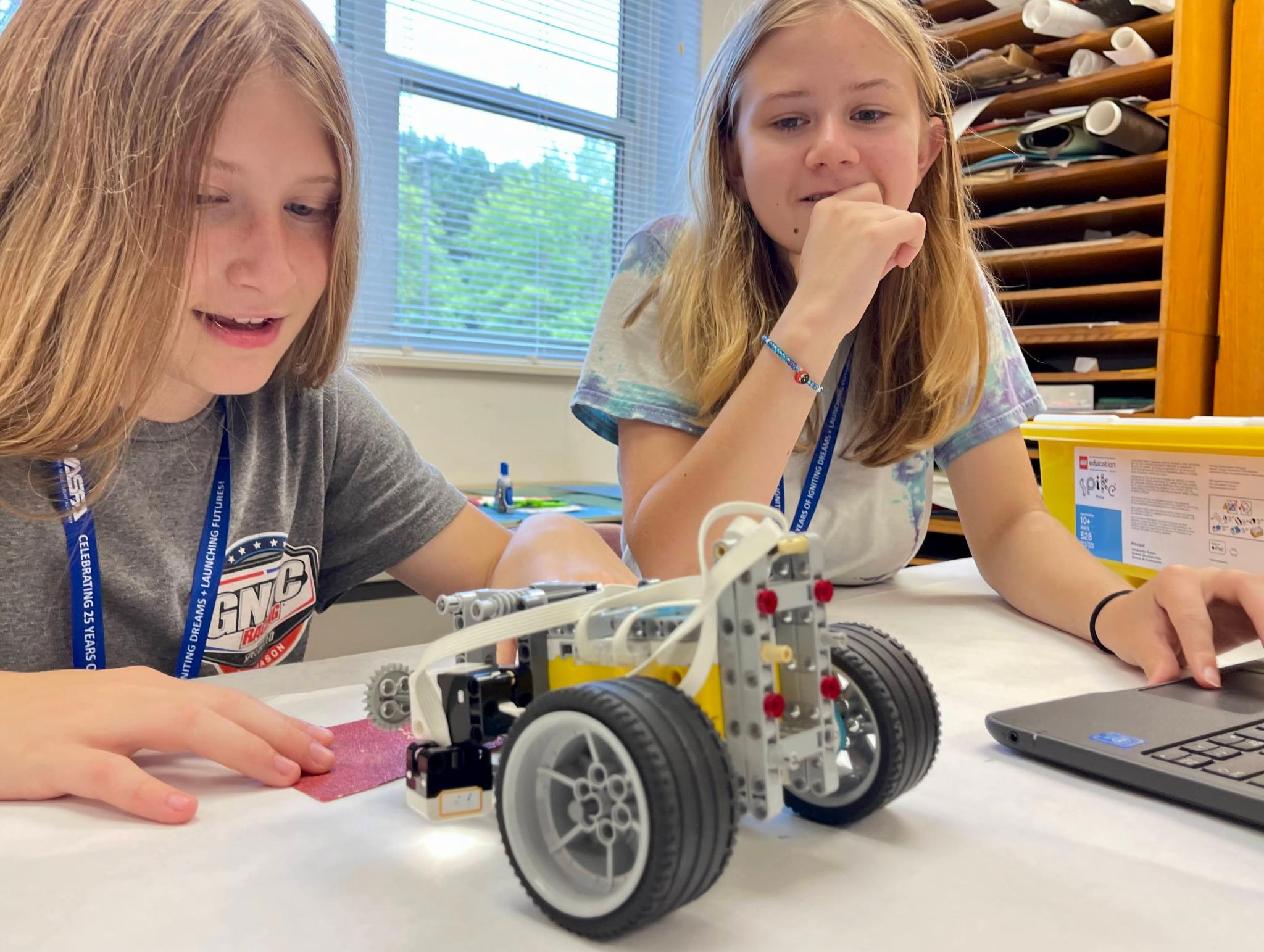 two young girls are looking at a small robot on a work table. The robot appears to be made of legos and has two black wheels