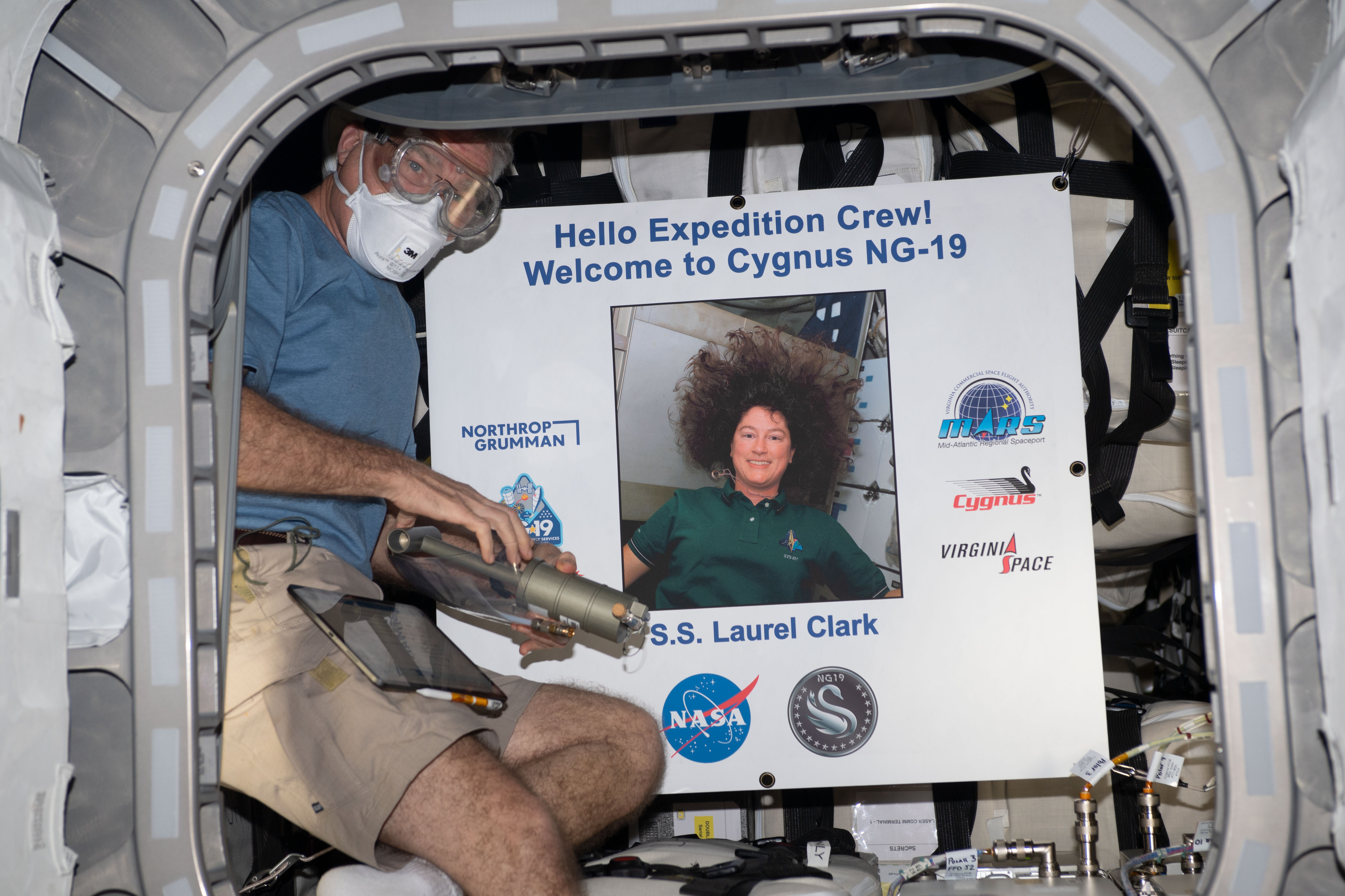 NASA astronaut and Expedition 69 Flight Engineer Stephen Bowen wears personal protective equipment (PPE) shortly after entering the Northrop Grumman Cygnus space freighter. The PPE protects an astronaut from eye contact with and breathing in any dust or debris that may have dislodged during the spacecraft's ascent to orbit. Bowen poses with a portrait of NASA astronaut Laurel Clark, whom the Cygnus cargo craft is named after, who perished aboard space shuttle Columbia during its return to Earth on Feb. 1, 2003.